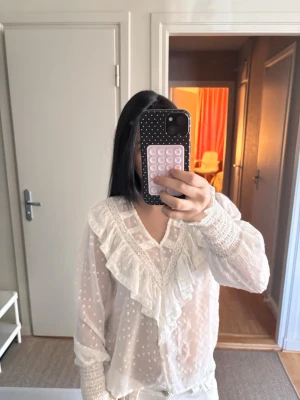 Vit blus med volanger och spetsdetaljer - Söt vit blus med prickigt mönster, volanger längs V-ringningen och spetsdetaljer. Blusen är genomskinlig och har knappar framtill samt långa ärmar med smock vid ärmsluten. Den har inga skador och använd fåtal gånger. Skriv gärna för mer.💕