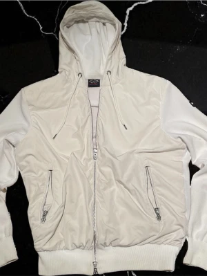 Vit cardigan från Paul & shark - Säljer en stilren vit pufferjacka med huva och dragsko. Jackan har två dragkedjeförsedda fickor framtill, ribbade muddar och quiltad insida. Materialet är en mix av glansig polyester och stickad bomull på ärmarna. Perfekt för kalla vinterdagar.