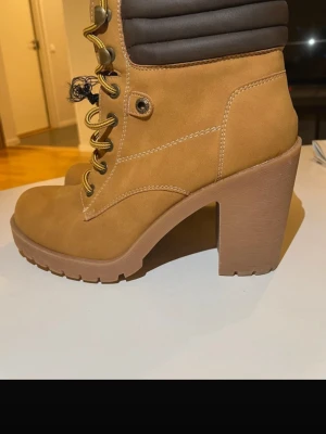 Gula stövletter med klack och snörning - Snygga stövletter i gul mockaimitation med kraftig klack och grov sula. Skorna har snörning framtill med guldfärgade öljetter och vadderad brun kant upptill för extra komfort. Perfekta för dig som vill ha en cool och trendig look.