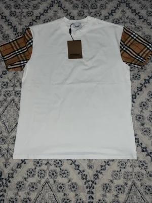Vit Burberry Helt ny - Säljer en clean vit t-shirt från Burberry med klassiska bruna, svarta och röda rutiga kortärmade ärmar. T-shirten har rund hals och är gjord i mjuk bomull. Perfekt för dig som vill ha en stilren look med en twist av ikoniskt mönster.