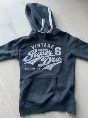 Svart hoodie från Superdry - Snygg svart hoodie från Superdry med vit vintage-tryck på bröstet och text i både engelska och japanska. Klassisk känguruficka framtill och vita dragsnören i huvan. Perfekt för en avslappnad streetwear-look.