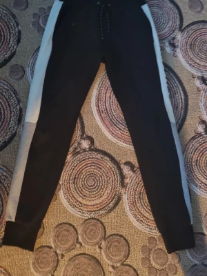 Svarta mjukisbyxor Jack & Jones - Snygga svarta mjukisbyxor från Jack & Jones med vita och grå sidopaneler. Byxorna har dragsko i midjan och tryckt logga längs ena benet. Perfekta för chill eller träning, med skön passform och mjukt material.