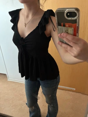 Svart volangtop med knyt - Säljer en svart topp med volangdetaljer vid axlarna och v-ringning. Toppen har knyt framtill och är lätt utsvängd nedtill för en flowy look. Perfekt till jeans eller kjol för en trendig outfit.