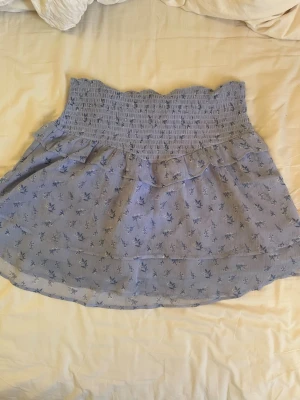 Kort ljusblå kjol med små blommor från Gina tricot - Kort skön kjol från Gina tricot i en ljusblåare färg med mörkblå blommor. Den går som omlott med volanger och är väldigt luftig och skön på sommaren. Den är från barnavdelning i storlek 170 men funkar för nästan vem som helst. 