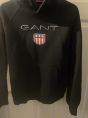 Svart hoodie från GANT med logga - Snygg svart hoodie från GANT med stor vit logga och ett broderat amerikanskt sköldmärke på bröstet. Klassisk huva och känguruficka framtill. Perfekt för en avslappnad och sportig stil. Tillverkad i mjukt material för extra komfort.