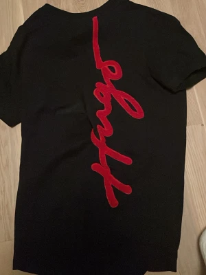 Svart t-shirt med röd logga från Hugo - Svart t-shirt från Hugo med en stor röd logotyp i snirklig stil längs ryggen. Klassisk passform och rund halsringning. Tillverkad i mjukt material som känns skönt mot huden. Perfekt för dig som gillar stilrena plagg med en twist.