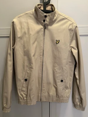 Beige jacka från Lyle & Scott - Stilren beige jacka från Lyle & Scott med dragkedja, hög krage och elastiska muddar. Jackan har två fickor med knappar och den klassiska gula fågel-loggan på bröstet. Insidan har rutigt foder för extra stilpoäng.
