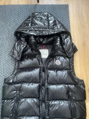 Svart dunväst från Moncler med huva - Snygg svart dunväst från Moncler med avtagbar huva och dragkedja framtill. Västen har två fickor med dragkedja och ikonisk Moncler-logga på bröstet. Tillverkad i glansigt material och fodrad med dun för extra värme. Perfekt för lager-på-lager-stil. Finns nfc scan och allting, tar dig direkt till moncler Hemsidan, skriv om ni har några funderingar! Med påse blir de 100kr extra