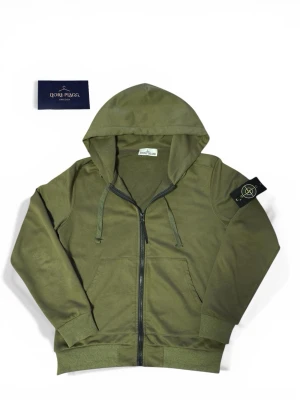 Stone Island Zip Hoodie - Snygg olivgrön hoodie från Stone Island med dragkedja framtill och klassisk logotyp-patch på vänster ärm. Huva med dragsko och ribbade muddar. Perfekt för en avslappnad streetwear-look.
