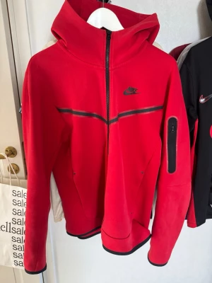 Röd Nike Tech Fleece hoodie - Nike Tech Fleece hoodie i rött med svarta detaljer och hel dragkedja. Hoodien har en huva, svarta linjer över bröstet och en praktisk ficka med dragkedja på ärmen. Perfekt för en sportig och stilren look.