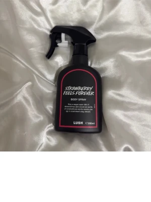 Lush Strawberry Feels Forever Body Spray - Strawberry Feels Forever Body Spray från Lush i en svart plastflaska med spraymunstycke. Doften har inslag av jordgubbar och vanilj, och flaskan rymmer 200 ml. Perfekt för dig som vill ha en söt och fräsch parfym i vardagen.