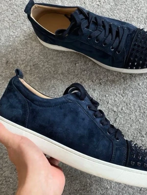 Mörkblå sneakers med nitar - Snygga mörkblå sneakers i mocka från Christian Louboutin med svarta nitar på tån och vita sulor. Skorna har svarta skosnören och en klassisk låg profil. Perfekta för dig som vill sticka ut med en edgy detalj.