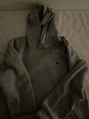 Grå hoodie från Lacoste - Snygg grå hoodie från Lacoste med klassisk krokodillogga på bröstet. Hoodien har en stor huva och är tillverkad i mjukt material som känns skönt mot huden. Perfekt för en avslappnad och stilren look.