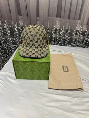 Gucci keps med GG-mönster - Snygg keps från Gucci i beige med klassiskt GG-monogram över hela kepsen. Kepsen har svarta detaljer och röd-gröna ränder på sidorna. Tillverkad i canvasmaterial och levereras med originalförpackning och dustbag. Äkta Keps kom privat