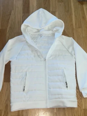 Vit pufferjacka med huva - Snygg vit pufferjacka med huva och dragkedja framtill. Jackan har två fickor med dragkedja och quiltad design på framsidan. Perfekt för kyligare dagar och enkel att matcha med olika outfits.