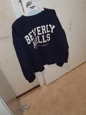 Svart Beverly Hills sweatshirt (Kvinna) - Cool svart sweatshirt med vit Beverly Hills-tryck på bröstet och texten 'Rodeo Drive'. Tröjan har rund hals, långa ärmar och ribbade muddar vid ärmslut och nederkant. Perfekt för en avslappnad och trendig look.