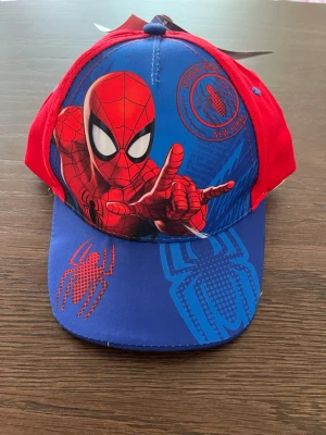 Spiderman keps i rött och blått - Cool keps med Spiderman-motiv från Marvel. Framsidan har en stor bild av Spiderman och spindeltryck, medan baksidan är helröd med blå detaljer. Justerbar passform och tillverkad i bomull. Perfekt för dig som gillar superhjältar och vill sticka ut.