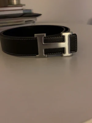 Svart skärp med H-spänne från H - Stilrent svart skärp från H med ett ikoniskt H-formad metallspänne. Skärpet är tillverkat i skinn med tydliga vita sömmar längs kanterna och har en klassisk, elegant design som passar till många outfits.