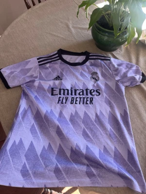 Lila Real Madrid matchtröja  - Snygg lila Real Madrid matchtröja från Adidas med svart krage och muddar. Tröjan har ett grafiskt mönster i olika lila nyanser, klubbmärke på bröstet och 'Emirates Fly Better' tryckt framtill. Perfekt för fotboll eller träning.