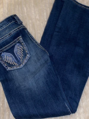 Mörkblå jeans med broderad ficka - Snygga mörkblå jeans med slitningar och broderad ficka med vinge i blått och guld. Klassisk femficksmodell med raka utsvängda ben och kontrastsömmar. Perfekta för dig som vill ha en unik detalj på dina jeans.
