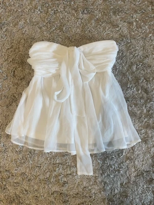 Vit bandeau topp med knytband - Supersöt vit bandeau topp i chiffongliknande material med knytband framtill som kan bäras både som rosett eller runt nacken. Toppens tyg är lätt och luftigt, perfekt för varma sommardagar. Den är från nelly i storlek 34 och har ett volangliknande fall. Använd 1 gång bara 