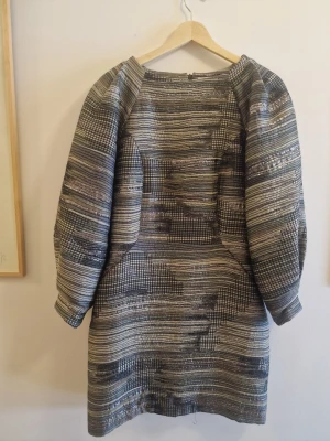 Mönstrad kort klänning i metallic från Rodebjer - Unik kort klänning med breda ärmar och ett coolt grafiskt vävt tyg i grått, svart och silver. Materialet har en lätt glansig metallic-effekt och klänningen har en figurnära passform med stora ärmar Perfekt för dig som gillar statement-plagg!