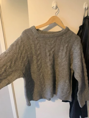 Grå kabelstickad tröja - Mysig grå stickad tröja med kabelstickat mönster och rund hals. Tröjan har en avslappnad passform och långa ärmar, perfekt för kyliga dagar. Materialet ser ut att vara mjukt och värmande, vilket gör den extra skön att bära.