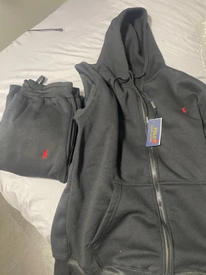 Svart loungewear-set från Polo Ralph Lauren - Svart loungewear-set från Polo Ralph Lauren med ärmlös hoodie och matchande byxor. Båda plaggen har det klassiska röda Polo-logot broderat. Hoodien har dragkedja och fickor framtill. Perfekt för chill dagar hemmaOBS helt ny me etiket köpt för o sälja vidare 