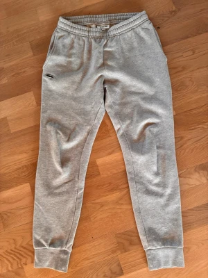 Gråa mjukisbyxor från Lacoste - Snygga grå mjukisbyxor från Lacoste med klassisk krokodillogga på vänster sida. Byxorna har resår i midjan, sidofickor och muddar vid bensluten. Perfekta för en avslappnad stil. Tillverkade i mjukt bomullsmaterial.