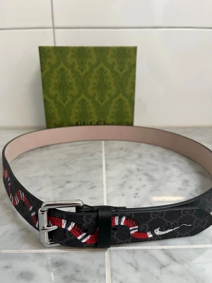 Gucci bälte med ormmönster - Snyggt bälte från Gucci med svart bas och ett iögonfallande ormmönster i rött, vitt och svart. Bältet har ett klassiskt spänne i silverfärgad metall och är tillverkat i skinn. Perfekt accessoar för att lyfta din outfit.