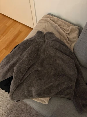 Grå teddykofta med halv dragkedja - Mysig grå teddy kofta med hög krage och halv dragkedja framtill. Jackan har en oversized passform och är gjord i fluffigt teddy-material som känns riktigt skönt. Perfekt för dig som vill ha en trendig och bekväm look under kyliga dagar.