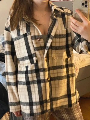 Beige och svart rutig overshirt - Snygg overshirt i beige med svart och grått rutmönster. Modellen har krage, knäppning framtill och två bröstfickor. Overshirten är i ett mjukt, lite tjockare tyg som passar perfekt till lager-på-lager-stil. Perfekt för dig som gillar en avslappnad och trendig look.