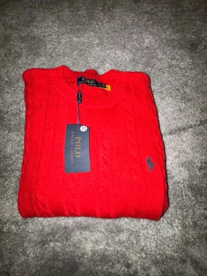 Röd  tröja Polo Ralph Lauren - Riktigt snygg röd kabelstickad tröja från Polo Ralph Lauren med rund halsringning och broderad blå logga på bröstet. Tröjan har långa ärmar och ribbade muddar. Perfekt för dig som vill ha en klassisk och stilren look.