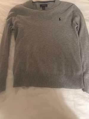 Grå tröja från Polo Ralph Lauren - Snygg grå tröja från Polo Ralph Lauren med klassisk rund hals och diskret broderad logga på bröstet. Tröjan är tillverkad i mjukt material och har ribbade muddar vid ärmslut och nederkant. Perfekt för en stilren look.