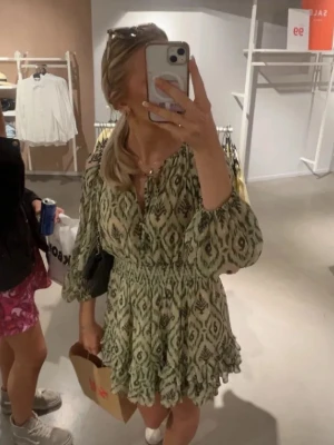 Grön mönstrad omlottklänning - Säljer en snygg grön kortklänning med grafiskt mönster och omlott-detalj. Klänningen har långa ärmar med resår vid handlederna och markerad midja. Perfekt för sommarens alla tillfällen!