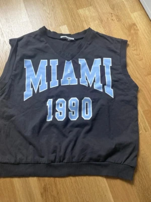 Svart väst med MIAMI-tryck - Cool svart väst med blått och vitt MIAMI 1990-tryck framtill. V-ringad modell med ribbad nederkant, perfekt för en avslappnad streetstyle-look. Tillverkad i mjuk bomull som känns skön mot huden. Det är för barn så storleken är 158.