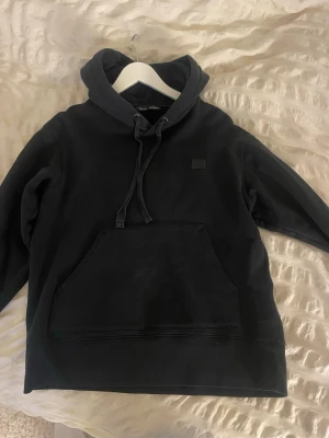 Svart hoodie från Acne Studios - Säljer en svart hoodie från Acne Studios med det klassiska Face-logotypen på bröstet. Hoodien har huva med dragsko och en stor magficka framtill. Tillverkad i mjuk bomull för skön känsla och avslappnad passform.