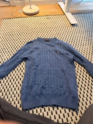 Blå kabelstickad tröja från Morris - Snygg blå kabelstickad tröja från Morris med rund halsringning och ribbade muddar. Tröjan har ett broderat logomärke på bröstet och är perfekt för dig som gillar klassisk stil. Passar till jeans eller chinos.