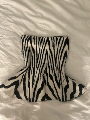 Zebramönstrad tubtopp i svart och vitt - Säljer en snygg zebramönstrad tubtopp i svart och vitt. Toppen har en utsvängd form nedtill och är axelbandslös, perfekt för dig som vill sticka ut. Materialet känns mjukt och stretchigt, vilket gör den bekväm att bära. Perfekt att matcha med jeans eller kjol.