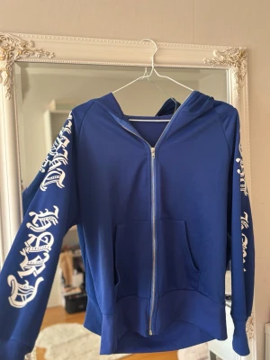 Blå hoodie med tryck på ärmarna - Snygg blå hoodie med dragkedja och vita tryck i gotisk stil längs båda ärmarna och huvan. Hoodien har känguruficka framtill och är tillverkad i mjukt material, perfekt för en avslappnad streetwear-look.