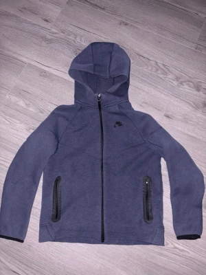 Nike tech tracksiut - Mörkblå mjukisbyxor från Nike med svarta detaljer och dragsko i midjan. Byxorna har ribbade muddar vid bensluten och en dragkedjeförsedd ficka på sidan. Perfekta för chill eller träning. Samma sak med tröjan också.