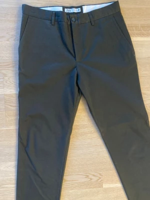 Mörkgröna chinos från Zara - Snygga mörkgröna chinos från Zara med klassisk rak passform och diskreta sidfickor. Byxorna har bälteshällor och stängs med knapp och dragkedja. Perfekta för en stilren och avslappnad look.
