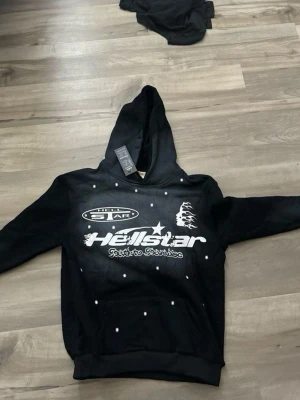 Svart Hellstar hoodie med tryck - Svart hoodie från Hellstar med vit logga och tryck framtill. Hoodien har huva och ribbade muddar vid ärmslut och nederkant. Snygga detaljer med små vita prickar över hela framsidan. Perfekt för en streetwear-look.