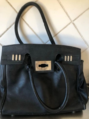 Svart handväska från Gina Tricot - Stilren svart handväska från Gina Tricot i fejkläder med två handtag och silverfärgade metalldetaljer. Väskan har ett klassiskt fyrkantigt snitt och innerfack med dragkedja. Perfekt för dig som gillar en enkel men trendig look.     Är välanvänd  men Håller ett tag till därav priset 