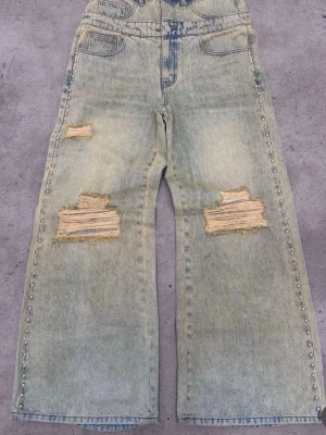 Ljusblå slitna jeans med vida ben - Coola ljusblå jeans med bred passform och slitningar på båda benen. Jeansen har råa hål och nitar längs sidorna för en edgy look. Perfekta för dig som gillar streetstyle och vill sticka ut. Klassisk femficksmodell i denim.