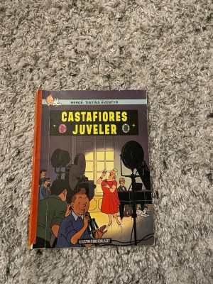 Castafiores juveler Tintins äventyr - Säljer seriealbumet 'Castafiores juveler' från Tintins äventyr av Hergé. Boken har hårda pärmar med färgglad framsida där Tintin och andra karaktärer syns i en studioscen. Ryggen är orange och boken är tryckt på papper.