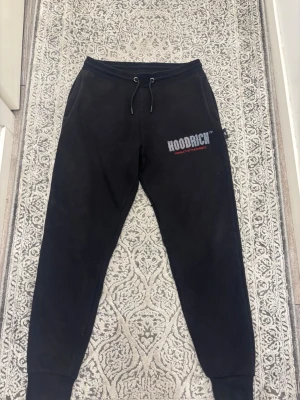 Svarta mjukisbyxor från Hoodrich - Svarta mjukisbyxor från Hoodrich med dragsko i midjan och tryckt logga på vänster lår. Byxorna har en avslappnad passform och muddar vid bensluten. Perfekta för en chill och street-inspirerad stil.