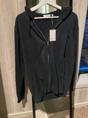Marinblå zip-hoodie från RYVLS - En stilren marinblå hoodie från RYVLS med dragkedja framtill, huva med dragsko och två fickor. Perfekt för en avslappnad look. Mjuk och skön känsla, passar till det mesta.