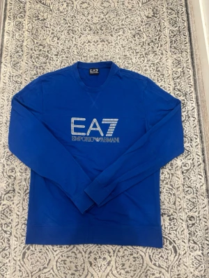 Blå EA7 Emporio Armani sweatshirt - Snygg blå sweatshirt från EA7 Emporio Armani med stort logotryck på bröstet. Tröjan har rund halsringning, långa ärmar och ribbade muddar. Perfekt för en avslappnad och trendig stil.