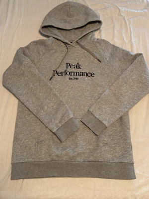Grå hoodie från Peak Performance - Snygg grå hoodie från Peak Performance med svart logga och text 'Est. 1986' på bröstet. Klassisk modell med huva och dragsnören, samt magficka framtill. Tillverkad i mjukt sweatshirtmaterial och har ribbade muddar vid ärmar och nederkant.